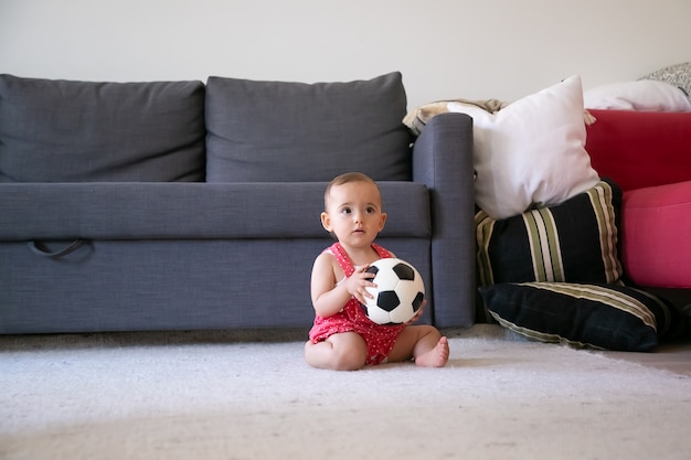 サッカーボールを持って、裸足でカーペットの上に座って、目をそらしている思いやりのある女の赤ちゃん。ソファの近くで自宅で遊んでいる赤いダンガリー
