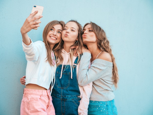 無料の写真 夏服の3人の若い笑顔の流行に敏感な女性 スマートフォンでselfieセルフポートレート写真を撮る女の子 壁の近くの通りでポーズ をとるモデル 肯定的な顔の感情を示す女性
