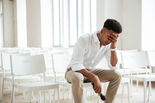 Tired african man sitting in office indoors holding microphone. Free Photo