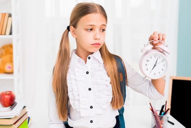 Tired schoolgirl with backpack holding clock in classroom Free Photo
