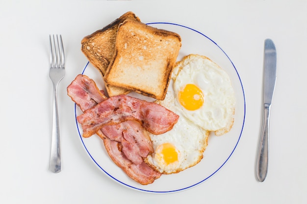 Toast breads; bacon and half fried eggs on ceramic plate with fork and butter knife isolated on white background Free Photo