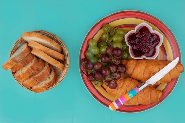 青い背景にクロワッサングレープラズベリージャムとナイフでパンのスライスを設定した朝食の上面図 無料の写真