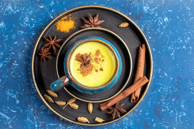 青の背景に伝統的なインドのアーユルヴェーダの黄金のターメリックミルクと食材のカップとプレートの平面図です プレミアム写真