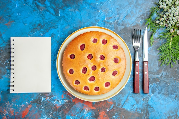 Top view of raspberry cake on oval plate cones fork knife a notebook on blue surface Free Photo
