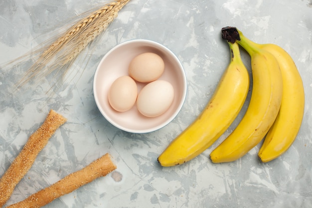 Top view yellow bananas with raw eggs and bun bread on light white desk Free Photo