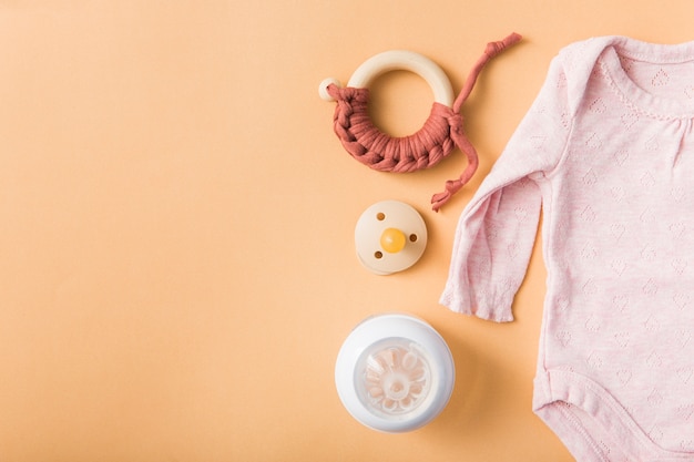 Toy; pacifier; milk bottle and pink baby onesie over an orange background Free Photo