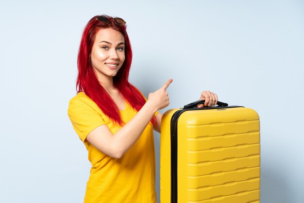 Traveler girl holding a suitcase isolated on blue pointing back ...