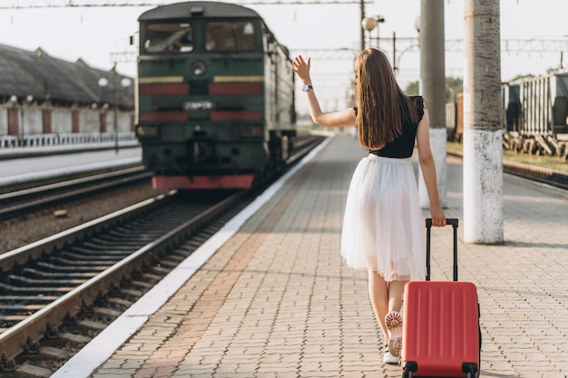 鉄道駅で電車を待っている白いスカートに赤いスーツケースを持つ旅行者 プレミアム写真