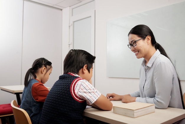 Two asian kids sitting in classroom, and smiling teacher in glasses talking to boy Free Photo