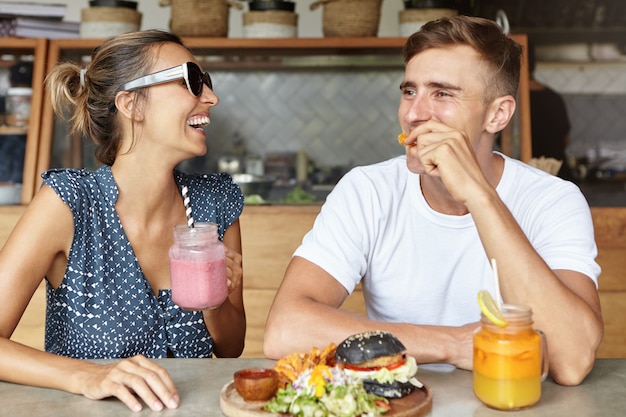 Two best friends having fun together and laughing while eating lunch at coffee shop. attractive female holding glass of pink smoothie enjoying lively conversation with her handsome boyfriend Free Photo