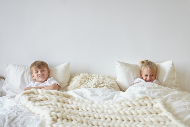 Two cute children relaxing in bedroom. indoor shot of teen boy in pajamas lying on bed with his blonde little brother on the other side, having playful looks. childhood, kids and family concept Free Photo