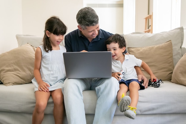 Two excited happy kids watching content on laptop with their dad while sitting on couch at home Free Photo