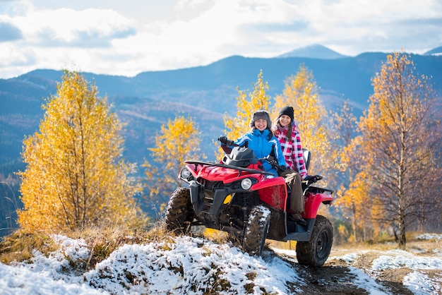 Two female riders atv in jackets and hats on a snow-covered trail at sunny autumn day against trees with yellow leaves and mountains Premium Photo