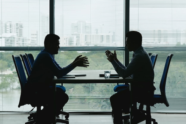 向かい合って​座って​交渉する​シャッター付き​の​オフィス​の​窓​に対する​2​つ​の​人間​の​輪郭 無料写真