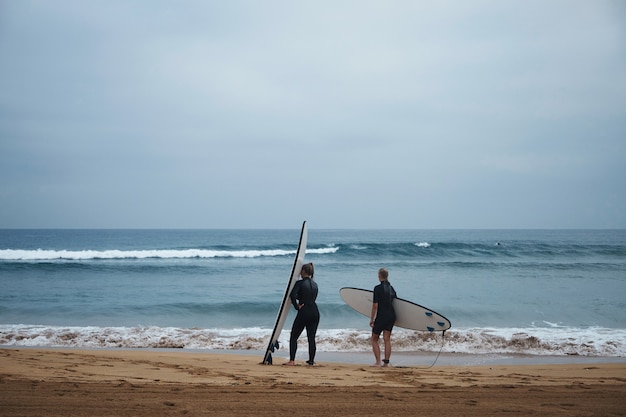 ロングボードを持った2人の認識できないサーフィンの女の子が海岸にとどまり 早朝に波を見て 完全なウェットスーツを着てサーフィンの準備ができています 無料の写真