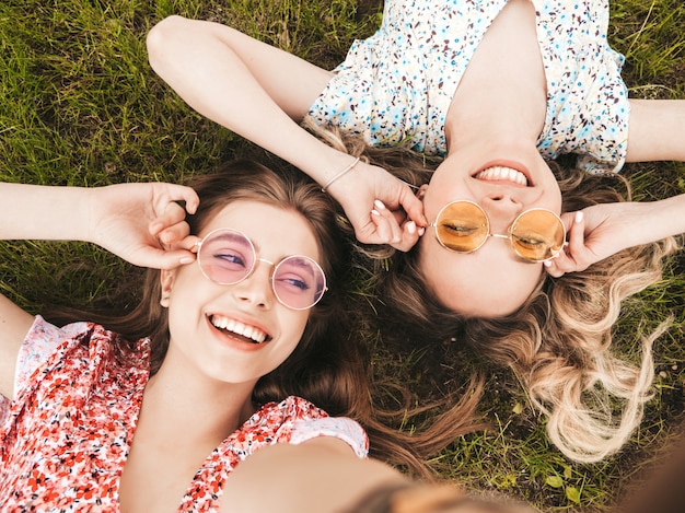 Two young beautiful smiling hipster girls in trendy summer sundress.sexy carefree women lying on the green grass in sunglasses.positive models having fun.top view.taking selfie photos on smartphone Free Photo