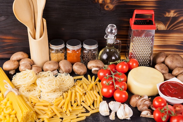 Uncooked raw macaroni, pasta and spaghetti next to fresh and healthy vegetables, variety of spices and sunflower oil on dark vintage rustic wooden background Free Photo