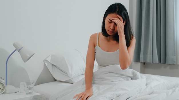 Unhealthy sick indian female suffers from insomnia. asian young woman taking painkiller medicine to relieve headache pain and drink glass of water sitting on bed in her bedroom at home in morning. Free Photo