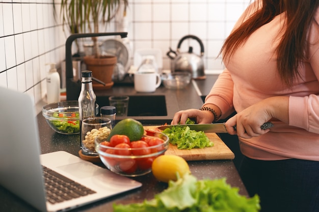 Unrecognizable plus size female standing at kitchen counter and cutting fresh organic vegetables on chopping board, making healthy low calories salad while watching movie online on laptop computer Free Photo