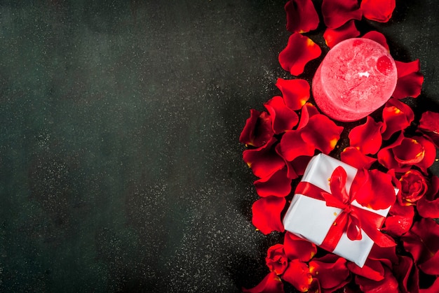 Valentine's day background with rose flower petals, white wrapped gift box with red ribbon and holiday red candle, on dark stone background, copy space top view Premium Photo