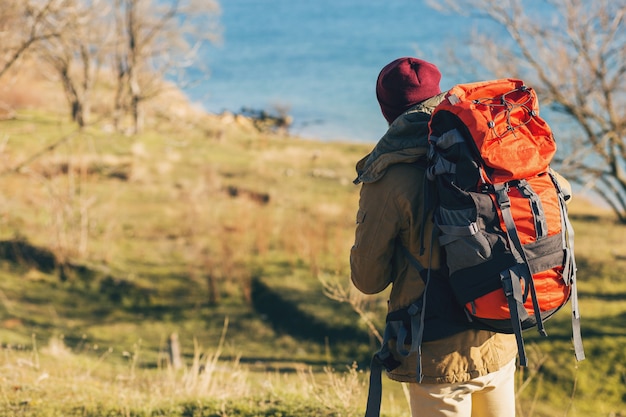 View from back on hipster man traveling with backpack wearing warm jacket and hat, active tourist, using mobile phone, exploring nature in cold season Free Photo