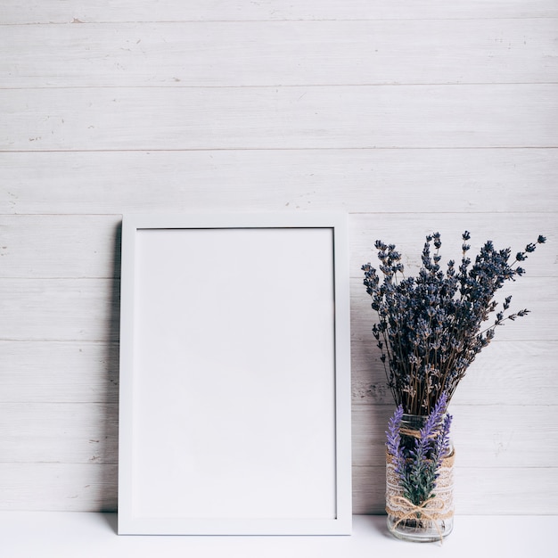 木製の背景の白い机の上のラベンダーガラス花瓶の近くの白い空白フレーム プレミアム写真