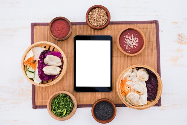 White blank screen digital tablet surround with sauce; chives and sesame seeds on placemat over texture backdrop Free Photo