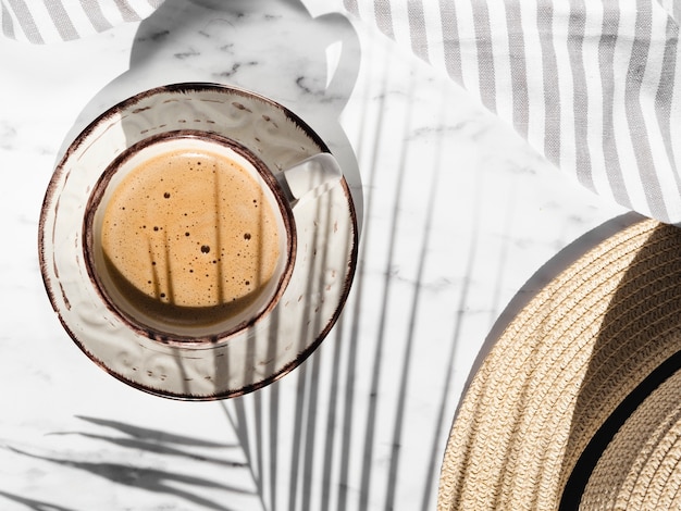 White cup with red shapes filled with creamy coffee on a white background with a striped grey and white cloth covered by a ficus leaf shadow and a hat Free Photo