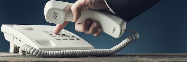  Wide view image of businessman dialing telephone number on white landline phone while holding a han