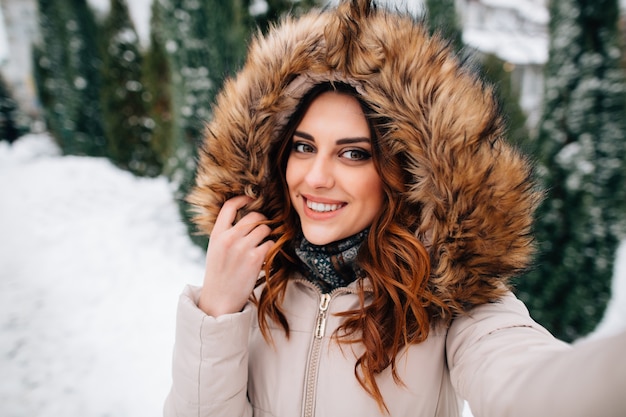 Winter selfie. Beautiful girl in fur hood takes selfie in snowy winter day Free Photo