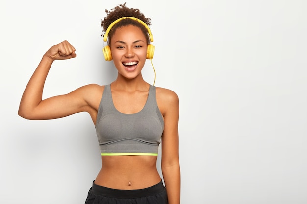 Woman in good mood, raises arm with muscles, has strong body, dressed in gym outfit, listens audio via modern headphones, poses indoor Free Photo
