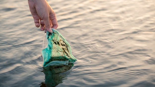 Woman hand picking up a discarded used disposable medical mask floats in sea waters. coronavirus plastic waste polluting the environment. trash in the beach threatening the health of oceans. Premium Photo