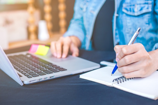 Woman hand works in a laptop computer and is writing on notepad ...