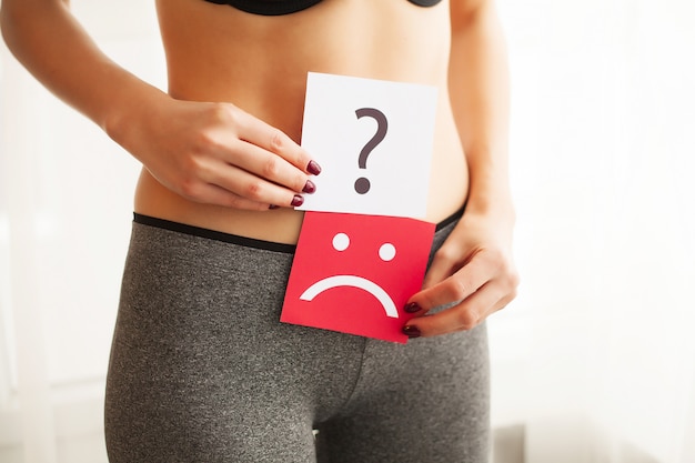 Woman health problem. closeup of female with fit slim body holding cards with sad smiley face and question mark near her stomach. digestive disorders, period pain, health issues concept Premium Photo