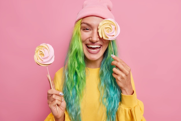 Free Photo Woman Holds Two Round Candies On Sticks Cons Eye With Delicious Caramel Lollipop Has Colorful Hair Wears Yellow Jumper And Hat Isolated On Pink