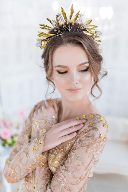 Woman in golden evening gawn and crown poses in luxury white room Free Photo