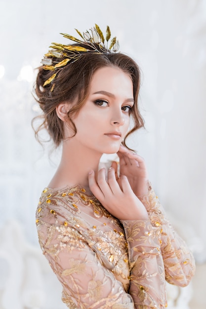 Woman in golden evening gawn and crown poses in luxury white room Free Photo