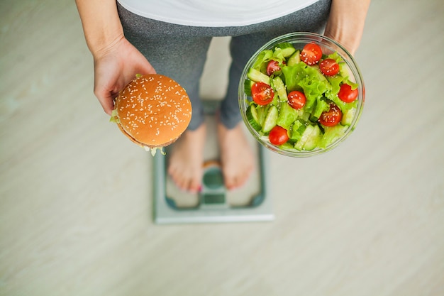 Woman Measuring Body Weight On Weighing Scale Holding Burger And Salad, Sweets Are Unhealthy Junk Fo