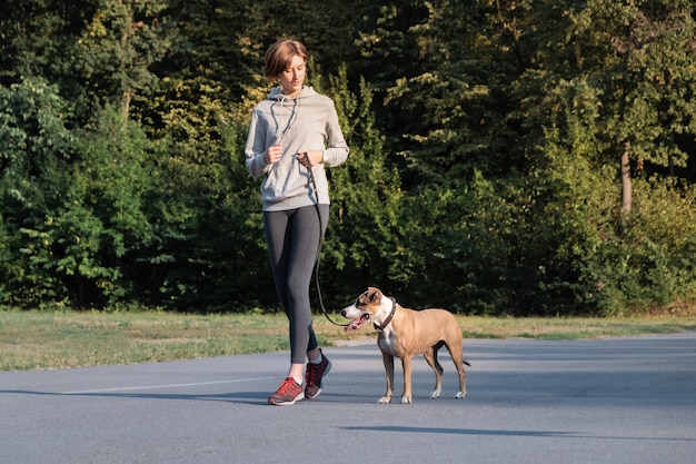 女性はジョギング中に走るように彼女の犬を訓練します ヤングは 身体運動をしている朝の散歩でメスとスタッフォードシャーテリア犬に合う プレミアム写真