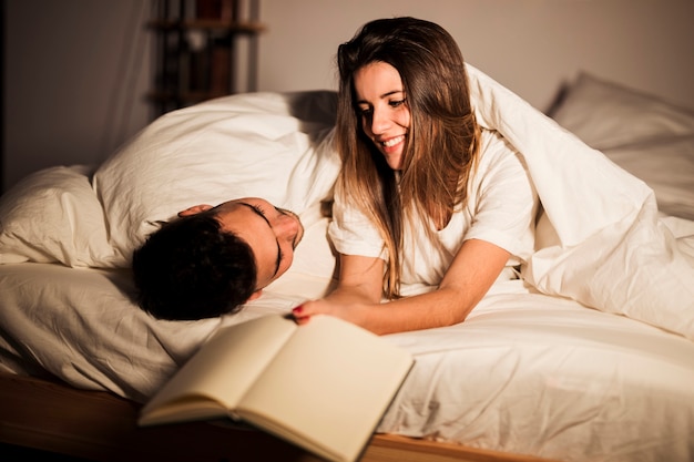 Woman With Book Near Man In Duvet Lying On Bed In Dark Room