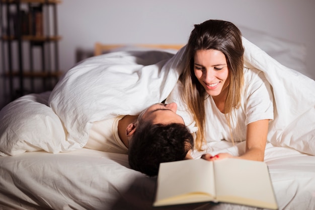 Woman With Book Near Man In Duvet Lying On Bed In Room Photo