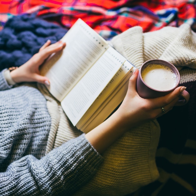 Woman with cup of coffee and book lies under colorful plaids Free Photo