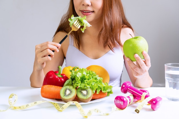 Premium Photo | Women eating vegetable while holding green apple with fruits,  vegetables, water, dumbbell and tape measure on white table. selection of  healthy food. clean eating and exercise concept.