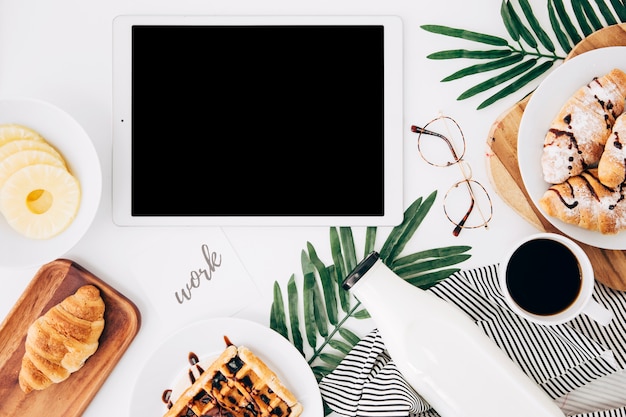 Work text on paper near the digital tablet; pineapple slices; croissant; waffles; bottle; coffee cup and eyeglasses on white desk Free Photo