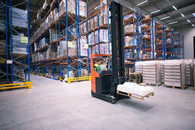 Worker operating forklift machine and relocating goods in large warehouse center Free Photo
