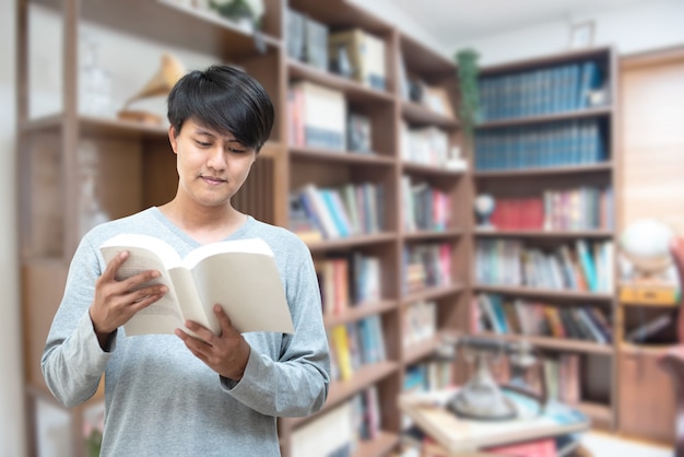 World book day concept.young asian man university student reading book sitting by bookshelf in college library for education research and self improvement. scholarship and educational opportunity. Premium Photo