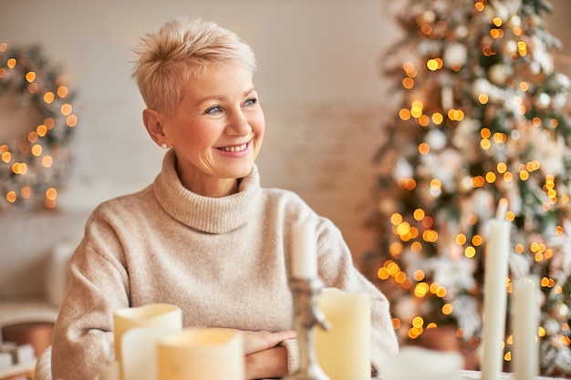 Xmas, holidays, decor, party and festive atmosphere concept. good looking cheerful middle aged female with short hair enjoying christmas mood, sitting around wax candles, decorations and lights Free Photo