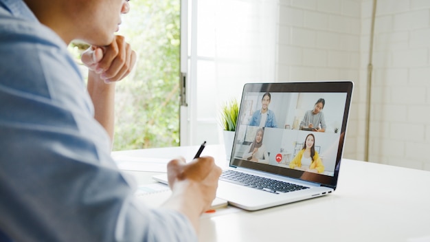 Young asia businessman using laptop talk to colleagues about plan in video call meeting while work from home at living room. self-isolation, social distancing, quarantine for corona virus prevention. Free Photo