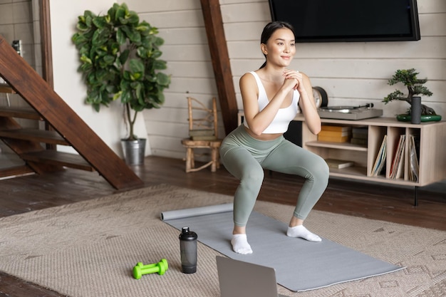 Young asian fitness girl doing squats workout at home follow online sport gym instructor standing on... Free Photo