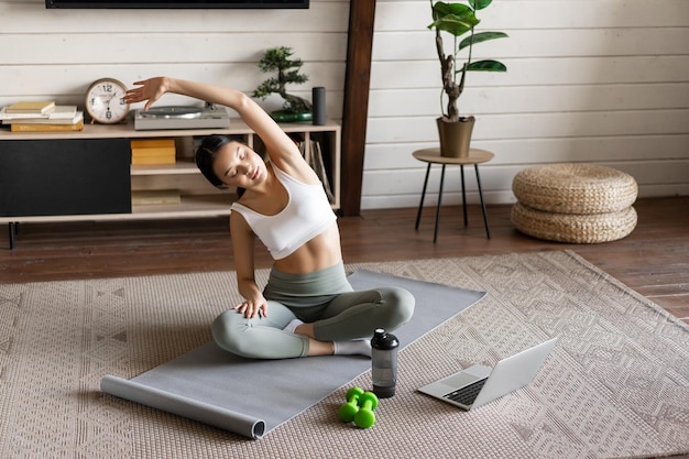 Young asian fitness girl doing stretching sport workout at home stretch arms and looking at laptop f... Free Photo
